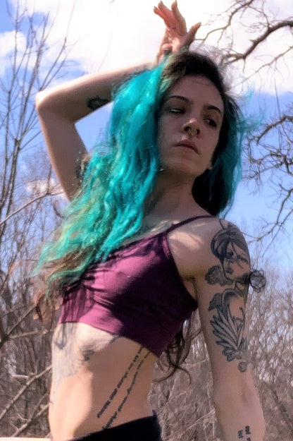 Side view of model with turquoise hair wearing a Plum Bliss Bralette, a purple bra with adjustable spaghetti straps and scrunched center puckering between the breasts. 90% cotton with 20% spandex.