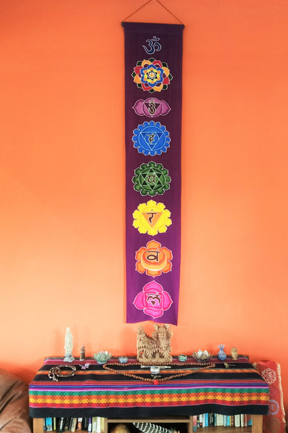 Vertical wall hanging with purple background and 7 rainbow chakra symbols. Handmade with wax and dye batik process on rayon fabric. Comes ready to hang. 