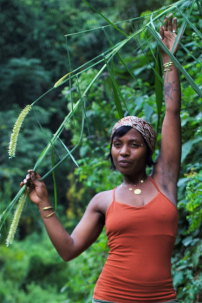 Womna with one hand up and one gripping plants wearing headscarf and gold jewelry with Rust Ananda Tank.