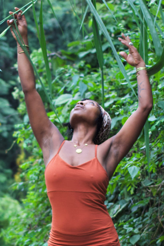 Woman with hands overhead in the jungle wearing Rust Ananda Tank. Shows seam under the breast area and full coverage with thin spaghetti straps. 